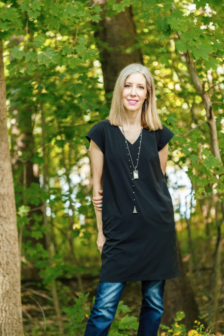 Blond-haired Stacey Wolf wearing a long black shirt, denim pants, and a loose necklace in the middle of the woods